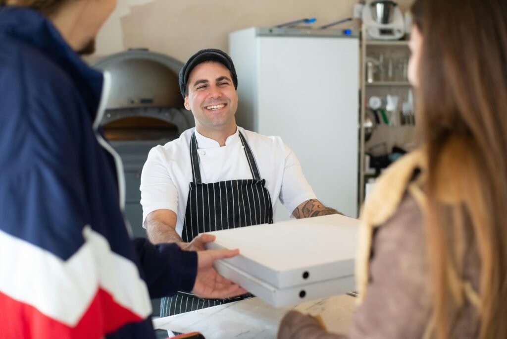 A cheerful chef handing pizza boxes to customers in a cozy Portuguese pizzeria.
